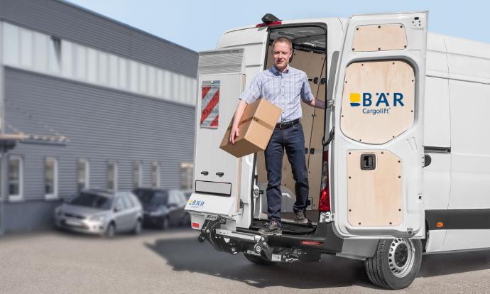 Kierowca wysiada z vana z paczką w ręku, mijając nieruchomą windę załadowczą BAR.