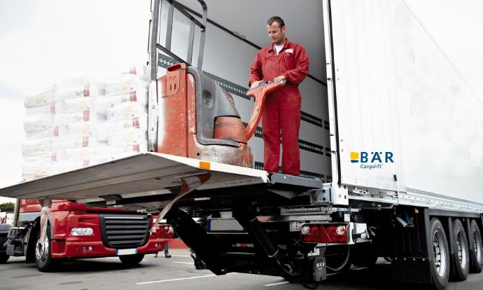 Kierowca z elektrycznym wózkiem paletowym i ciężką paletą na platformie windy załadowczej Bar