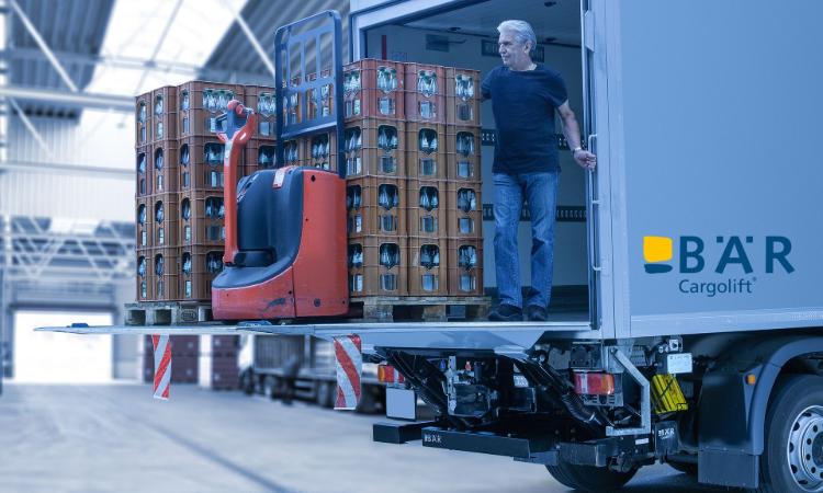 Kierowca stoi z kilkoma paletami z napojami i elektrycznym wózkiem paletowym na platformie windy załadowczej BAR
